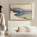 A person in a white robe stands in a minimalist room, gazing at the Winter Reflection Canvas Art - Horizontal, a framed painting of snowy mountains and a calm lake, hanging above a beige couch.