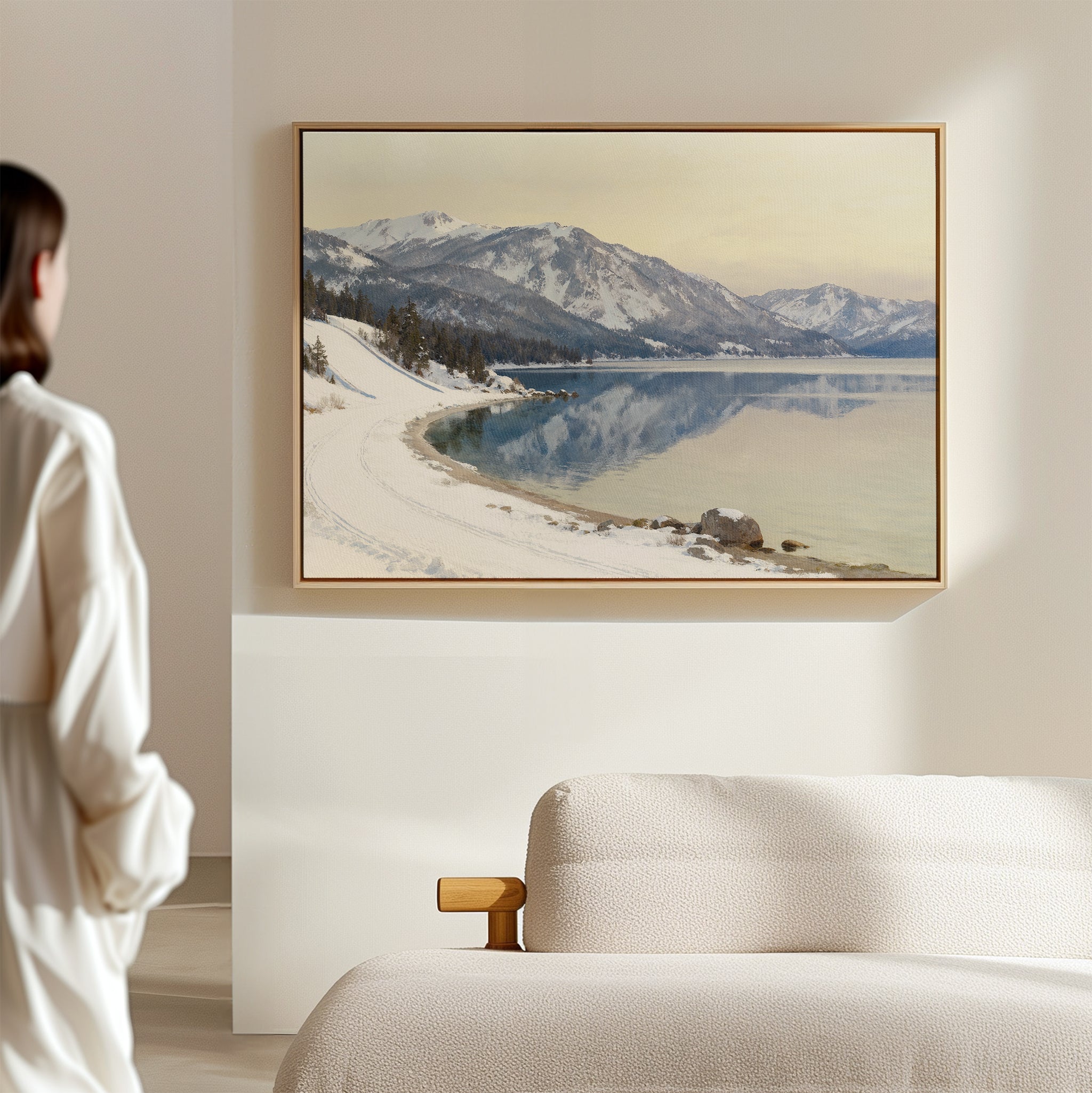 A person in a white robe stands in a minimalist room, gazing at the Winter Reflection Canvas Art - Horizontal, a framed painting of snowy mountains and a calm lake, hanging above a beige couch.