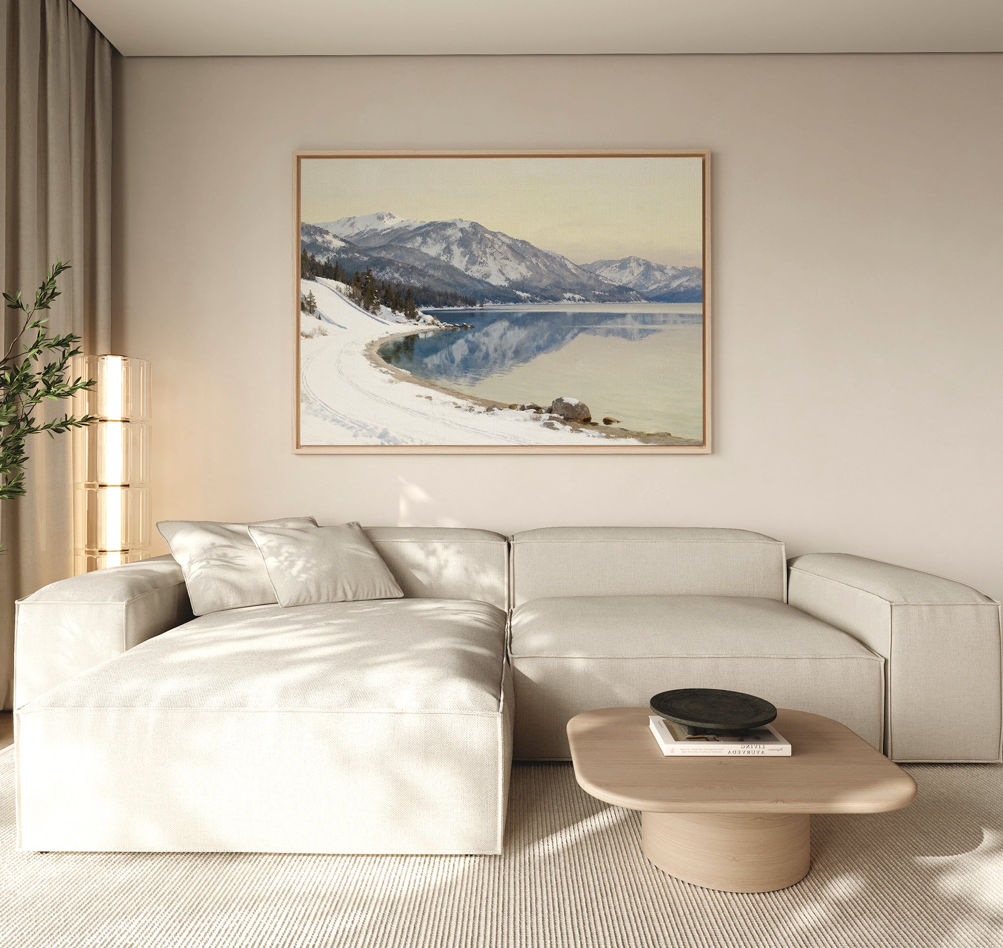 A modern living room with a light beige sectional, wooden coffee table, and serene decor featuring the Winter Reflection Canvas Art - Horizontal of snowy mountains and a lake displayed above the sofa.