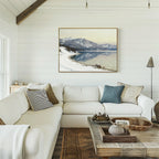 A cozy living room with a white sectional sofa, rustic wooden coffee table, and neutral decor. The white shiplap wall displays the Winter Reflection Canvas Art - Horizontal, showing a snowy alpine lake and mountain landscape.