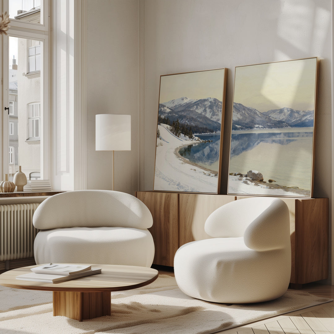 A modern living room with two rounded white chairs, a low wooden coffee table, and a wooden sideboard topped with the Winter Reflection - Set of 2 alpine lake wall art, all bathed in natural light from tall windows.