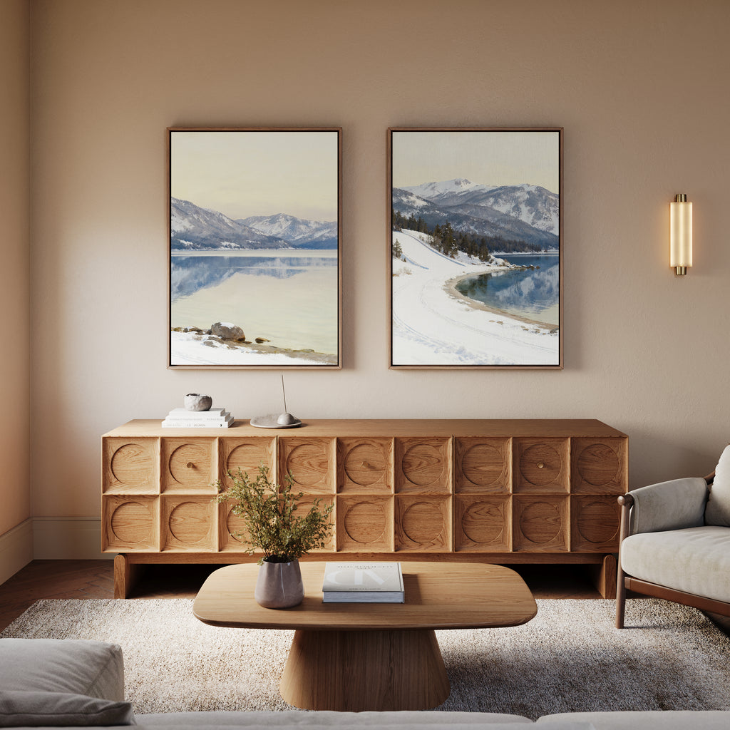 A cozy living room featuring a wooden sideboard, round wooden coffee table, vase with greenery, light sofa, and "Winter Reflection - Set of 2" alpine lake wall art depicting mountains and lakes on a beige wall.