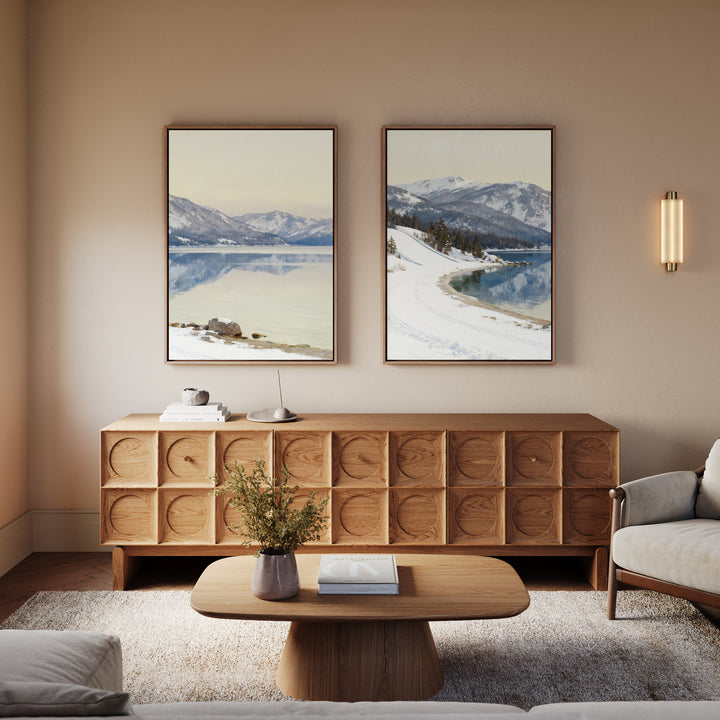 A cozy living room featuring a wooden sideboard, round wooden coffee table, vase with greenery, light sofa, and "Winter Reflection - Set of 2" alpine lake wall art depicting mountains and lakes on a beige wall.