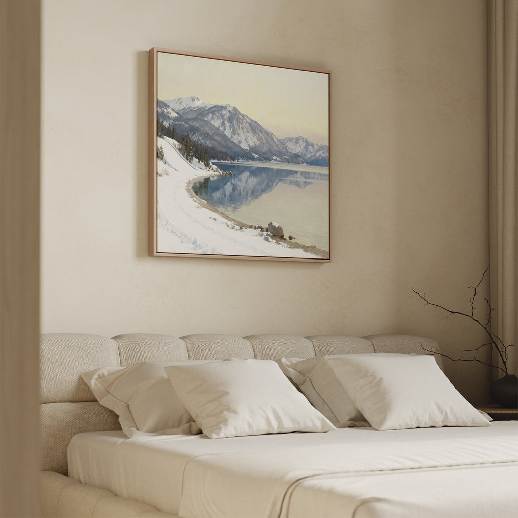A cozy beige bedroom features a neatly made bed, white pillows, and the Winter Reflection Canvas Art - Square—an alpine lake and snowy mountains print—above the headboard. A minimalist vase with bare branches sits in the corner.
