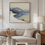 A cozy living room with a beige sofa, neutral cushions, lamp, and wooden side table. Above the sofa hangs the Winter Reflection Canvas Art - Square showing snowy mountains over a tranquil lake. Dried flowers sit on the coffee table.