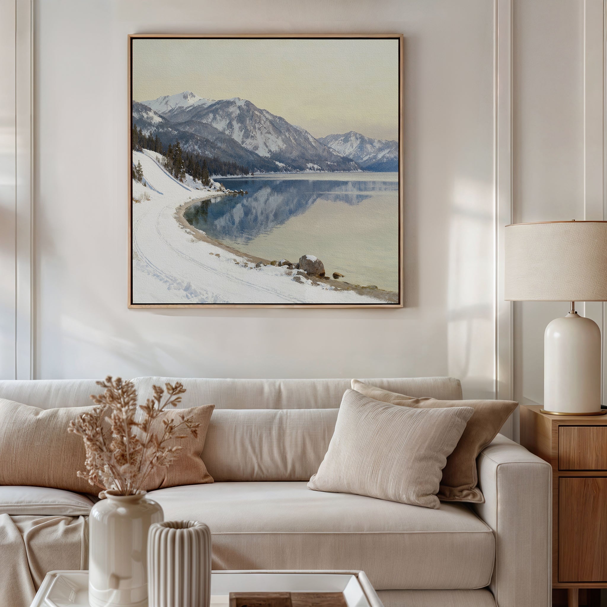 A cozy living room with a beige sofa, neutral cushions, lamp, and wooden side table. Above the sofa hangs the Winter Reflection Canvas Art - Square showing snowy mountains over a tranquil lake. Dried flowers sit on the coffee table.