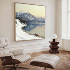 A modern living room features a white lounge chair and ottoman, a dark wood side table with a white lamp, and the Winter Reflection Canvas Art - Square, showcasing a snowy alpine lake with mirrored mountains.