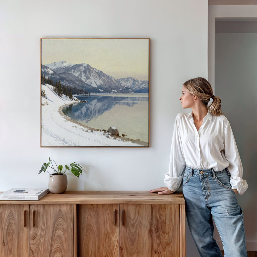 A woman in a white shirt and blue jeans stands in a modern room, leaning on a wooden sideboard and admiring the Winter Reflection Canvas Art - Square, which features mountains, snowy shores, and a calm alpine lake—a perfect winter scene.