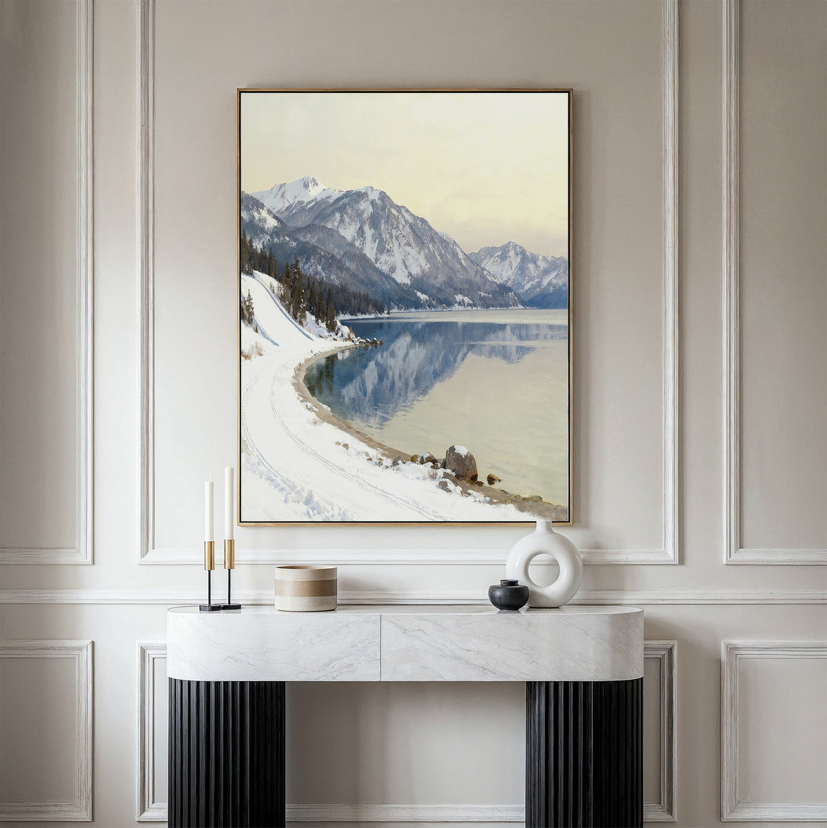A minimalist console table with decor stands against a light wall, topped by the Winter Reflection Canvas Art - Vertical, featuring snowy mountains and a tranquil alpine lake in winter.