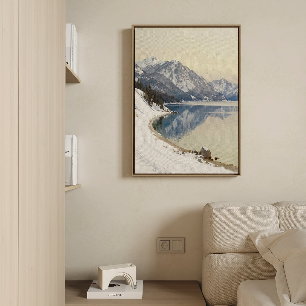 The Winter Reflection Canvas Art - Vertical, depicting snowy mountains and a calm lake, hangs on a beige wall above a cream sofa. To the left, a light wood bookshelf with books and white decor accentuates the tranquil ambiance.