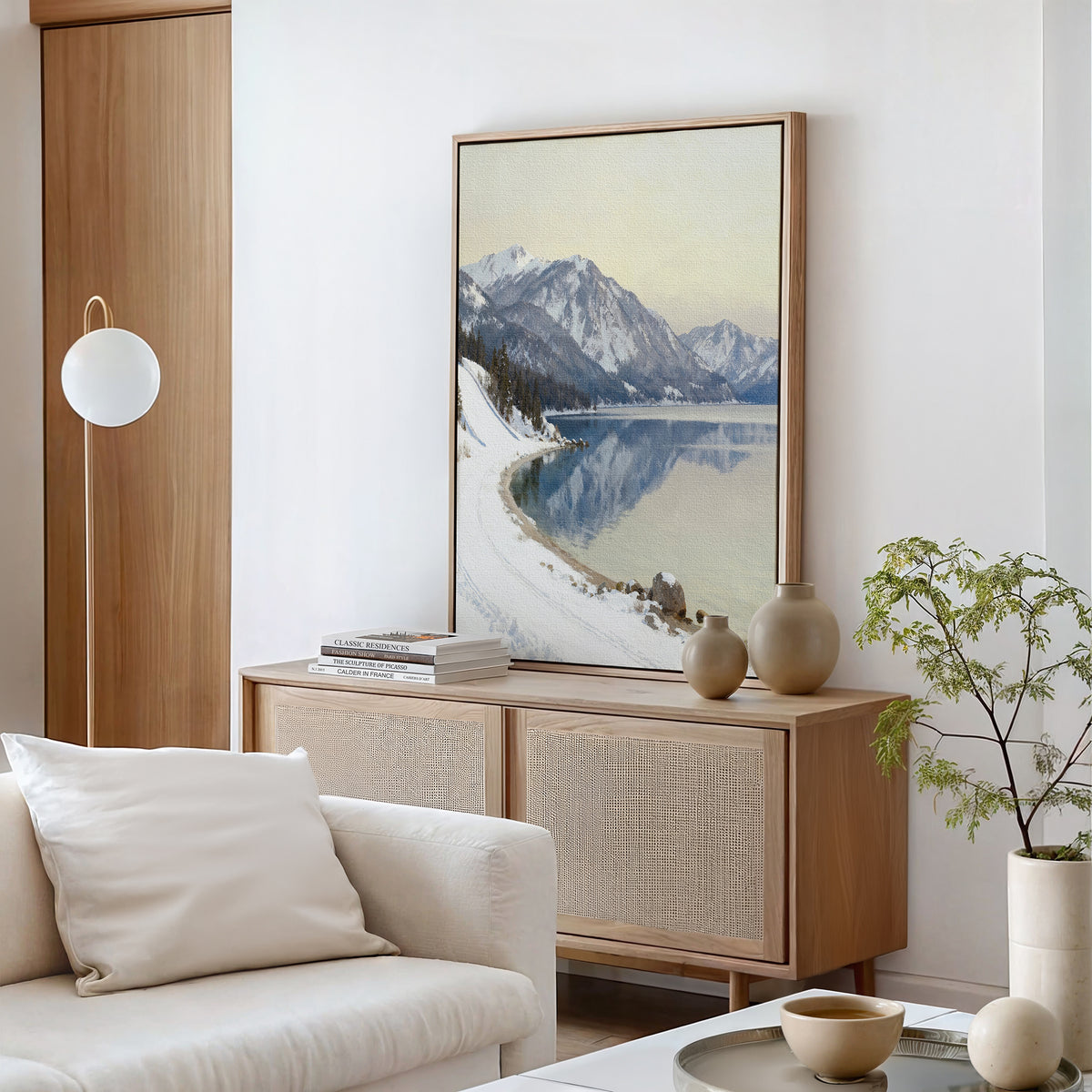 A minimalist living room featuring a light wood sideboard, neutral vases, the Winter Reflection Canvas Art - Vertical, a white sofa with cushions, a round coffee table, and a potted plant.