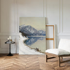 The Winter Reflection Canvas Art - Vertical, depicting a snowy mountain landscape, leans against a white-paneled wall beside an empty frame, with a cushioned wooden chair and black round table with candles on herringbone wood floors nearby.