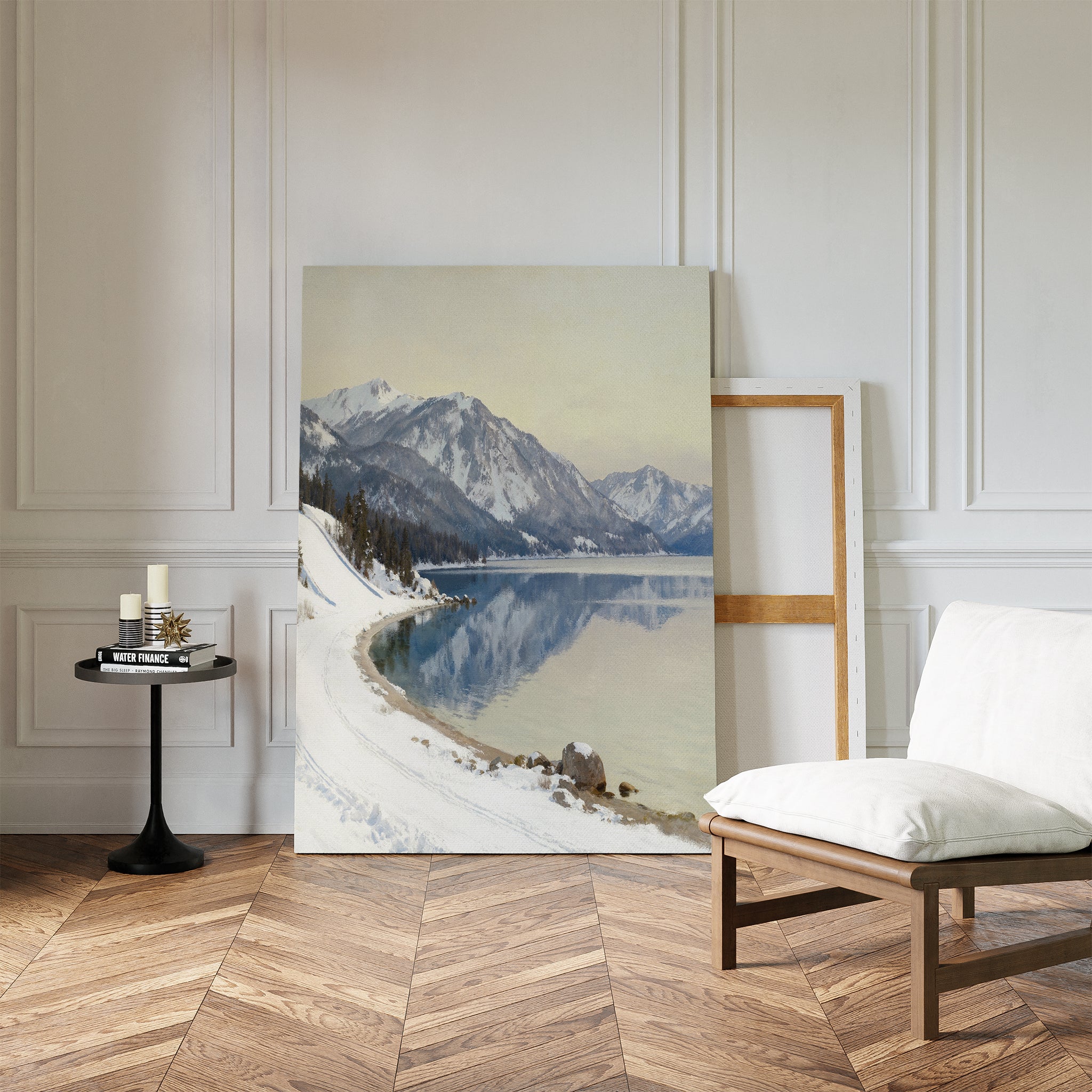 The Winter Reflection Canvas Art - Vertical, depicting a snowy mountain landscape, leans against a white-paneled wall beside an empty frame, with a cushioned wooden chair and black round table with candles on herringbone wood floors nearby.