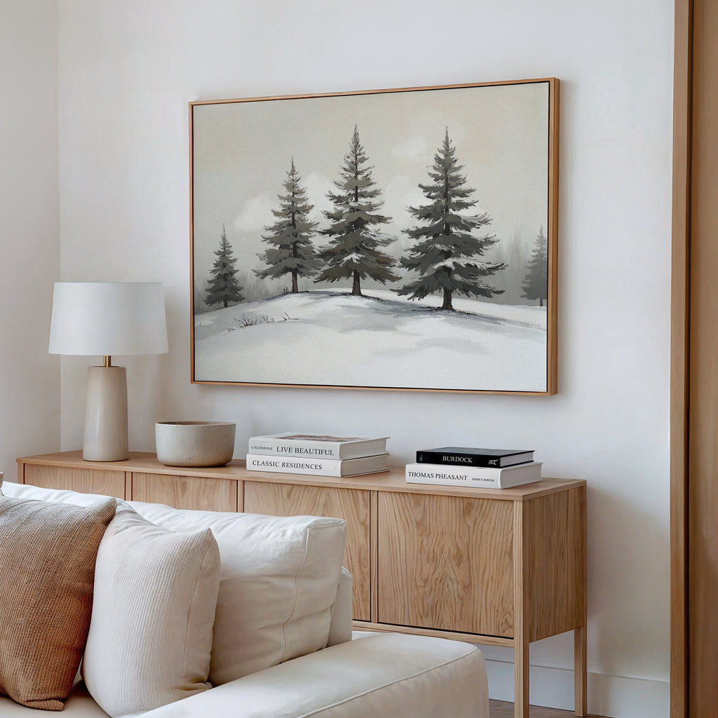 A cozy living room features a light wood sideboard with stacked books, a white lamp, and a bowl. Above hangs the Winter Solitude Canvas Art - Horizontal depicting snowy pine trees. A white sofa is partially visible in the inviting foreground.