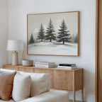 A cozy living room features a light wood sideboard with stacked books, a white lamp, and a bowl. Above hangs the Winter Solitude Canvas Art - Horizontal depicting snowy pine trees. A white sofa is partially visible in the inviting foreground.