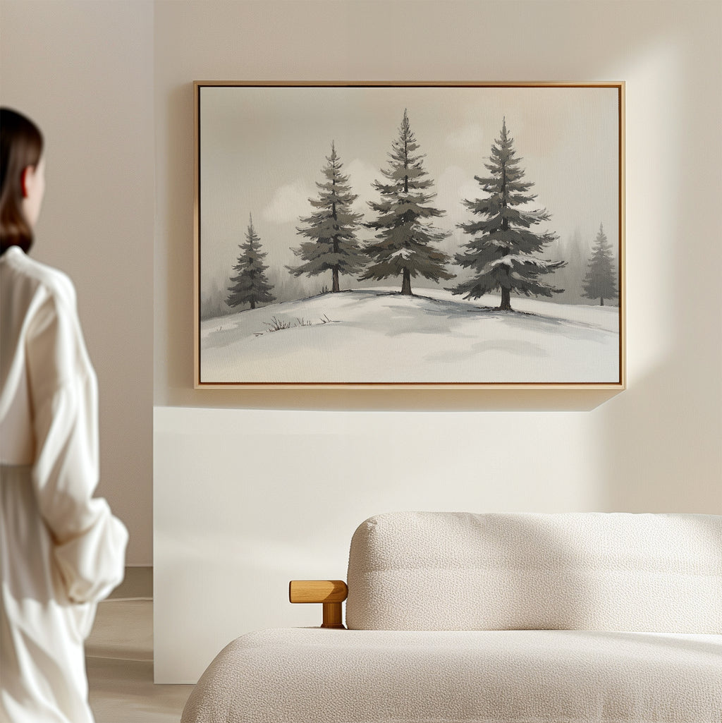 In a minimalistic room, a person admires the Winter Solitude Canvas Art - Horizontal, featuring snowy pine trees, hanging above a beige sofa on a light-colored wall.