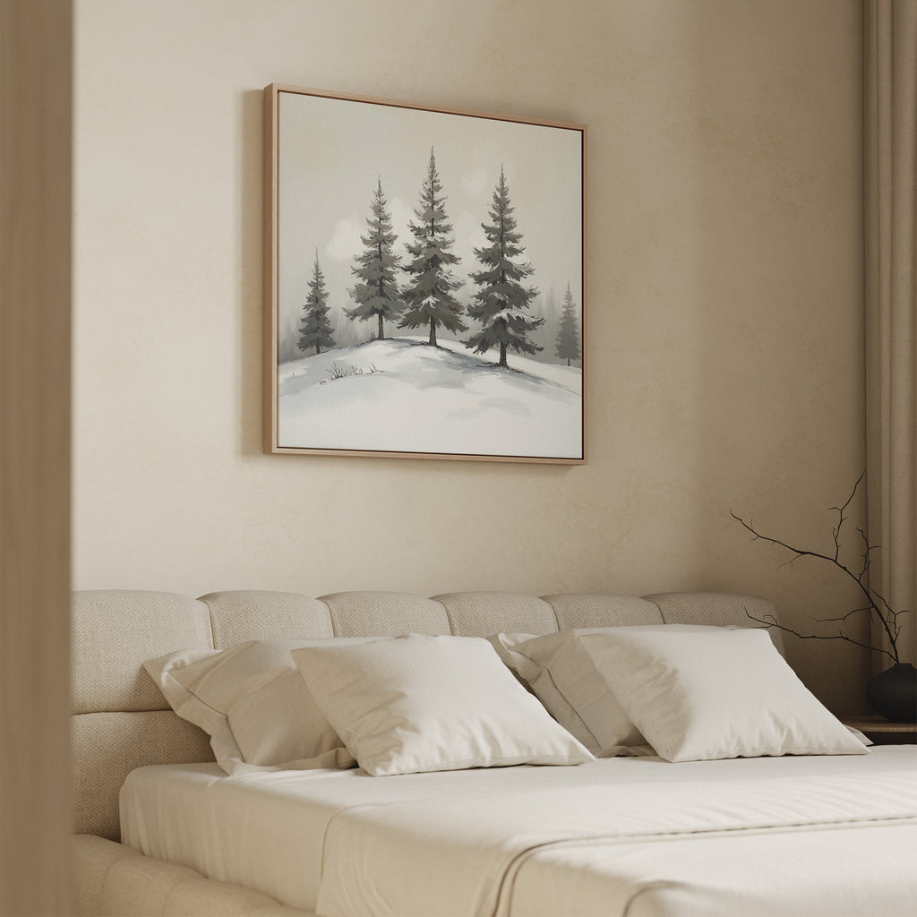 A cozy bedroom features a beige upholstered bed with white bedding and soft pillows. Above the bed hangs the Winter Solitude Canvas Art - Square, depicting pine trees in snow. A bare branch decorates a table in the corner.