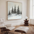 A modern living room with wood floors, a white lounge chair and ottoman, a dark side table with a small white lamp, and the Winter Solitude Canvas Art - Square featuring snowy pine trees displayed on the wall.