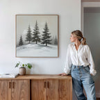 A woman in a white blouse and jeans stands by a wooden console with a small plant and books, admiring the Winter Solitude Canvas Art - Square featuring pine trees on a white wall.