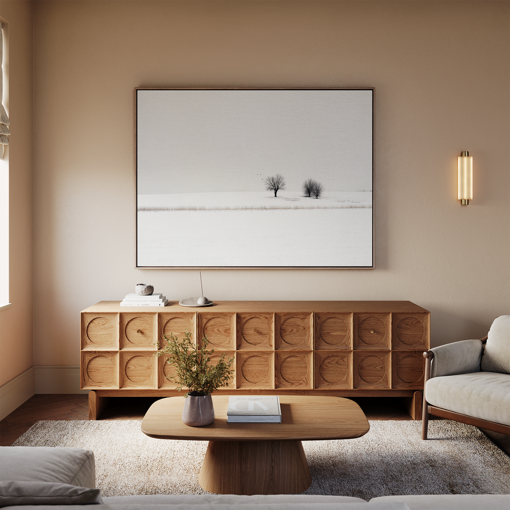 A cozy living room with beige walls, light sofas, a wooden coffee table, and a textured wooden sideboard. Above the sideboard hangs the Winter Silence Canvas Art, featuring two trees in snow—perfect for elegant winter decor.