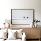 A minimalist living room features a light wood sideboard with ceramic vases, a lamp, a leafy plant, and the Winter Silence Canvas Art above. In front, a white sofa with pillows completes the serene look.